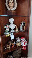 Overall view of wooden cabinet shelf with various porcelain and ceramic figurines including busts, salt and pepper shakers, clown, carousel music box, and Norman Rockwell style figures