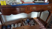 Front view of the Drexel sofa table showing two drawers with brass hardware and visible wear on the surface and legs. Cardboard boxes and mugs below are not included.