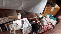 Assorted folded linens including Christmas-themed towels, checkered linens, and napkins with floral embroidery stacked on a surface with packaging tags visible.