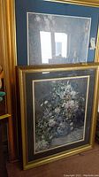 Two framed prints leaning against wall. One Monet print with blue matte border and gilded frame, larger size. Other floral print with dark matte and gilded frame, smaller size.