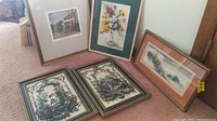 Three framed prints under glass arranged on carpeted floor showing variety of sizes and subjects including floral watercolor and classical etchings.