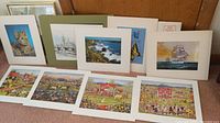 Nine unframed art prints laid out on floor, with various subjects including cat, ships, coastal scenes, butterfly, and folk art community scenes.