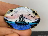 Front view of the oval brooch showing detailed enamel Viking ship scene with gold overlay and scalloped edges.