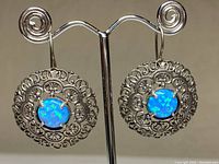 Front view of pair of sterling silver filigree earrings with blue-green opal centers on a metal display stand.