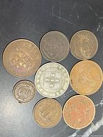 Top-down view of 8 assorted coins showing varied designs including ships, heraldic crests, and numeric denominations on dark surface.