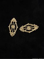 Two antique brass diamond shaped findings with deco style design shown individually on black background.