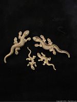 Four brass gecko figures displayed on black background showing details and sizes.