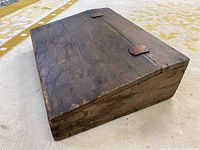 View of the angled antique wooden desk top, closed, showing the hinged lid and metal clasps with natural wood texture and signs of wear.