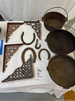 Full view of three iron cooking pots with handles, two large ornate iron shelf brackets, and four iron horseshoes on a white cloth.
