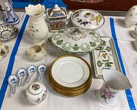 Full view of assorted china on a white tablecloth with blue border, including plates, vases, spoons, serving dishes and teapot.