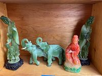 Shelf display of four resin statues: two tall green figures on black bases, one red-orange and green seated figure, two green elephants, one missing tusks