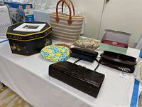 Photo showing a group of assorted handbags, wallets, pouches, a striped beach bag, and a Dobbs New York hatbox on a white table.
