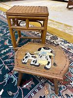 Photo of rectangular rattan foot stool and small wooden stool with cow painting on top, placed on decorative rug.
