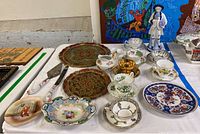 Assorted teacups, ornate collector's plates with red and gold designs, porcelain figurine, and various small dishes arranged on table