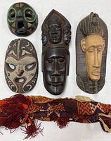 Four carved tribal masks and one textile camel accessory laid flat on white fabric. Various colors and paint patterns visible; masks are wooden with some painted decorations.