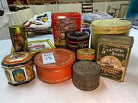Photo showing all 10 vintage tin cans arranged on white surface. Including Arnott's Venezia red biscuit tin, The Steamship Brand candy tin, Granger tin, Haeberlein-Metzger tin, and others.