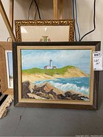 Two framed oil paintings showing lighthouse coastal scenes with rocks and ocean waves, stacked on floor with a glimpse of a third gold framed piece behind.