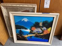Three framed paintings, two with glass, stacked leaning against a wall; showing one Japanese landscape painting with blue sky and mountain.