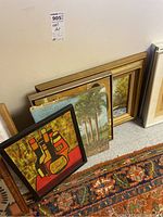 Multiple framed paintings stacked together showing edges, frames, and some visible art styles including abstract liquor bottles and landscape.