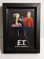 Frontal view of the black framed E.T. movie film strip collectible showing the photographic print of E.T. and a child, the film strip piece, and the E.T. logo beneath it.