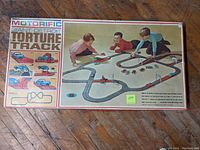 Full box top of Motorific Giant Detroit Torture Track slot car set with children playing on the track, showing the complete track layout and several slot cars in play.