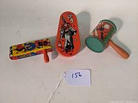 Three vintage lithographed tin hand noisemakers shown from front and sides on a flat surface. One with orange witch design, one with green drum witch design, one with colorful party dancing figures.