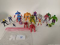 Group shot of 16 Marvel Comics action figures including Captain America, The Thing, Hulk, Iron Man, Ghost Rider, and others arranged standing and lying in front of white background with plastic bag in foreground.