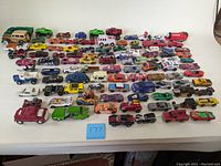 Overview photo showing about 60 assorted toy cars on a white cloth background; varies in colors and designs including some Red Line Hot Wheels and other collectible brands.