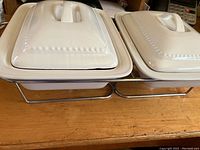 Two white rectangular ceramic dishes with lids resting on a metal frame designed for heating using tea lights.