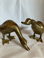 Close-up photo of two solid brass duck sculptures on white background, showing one duck bending down and the other standing upright.