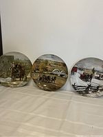 Three collector plates arranged horizontally showing rural scenes with horse carriages and farmhouses on each plate.