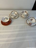 Four Aynsley porcelain tea cups and matching saucers arranged on a table with a white cloth. Variations in floral patterns and gold trim are visible.