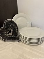 Stack of eight extra large white plates next to a dark gray metal heart-shaped cake pan with a smaller heart cutout in the center