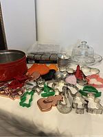 Photo showing red tarnished tin can with 29 assorted metal cookie cutters, set of metal letter presses, glass cheese plate with dome, and white serving plate on table.