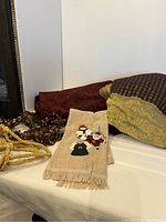 Photo showing two burgundy velvet tablecloths folded, partially visible gold and burgundy tablecloth, embroidered Christmas hand towel with snowman, Santa, and angel, and gold garland pieces coiled