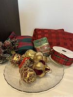 Six gold and burgundy Christmas ornaments, two types of red plaid ribbon, a folded plaid tablecloth, and red chair covers placed around a clear glass plate.