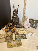 Photo showing grapevine rabbit sculpture, stuffed fabric rabbit doll, metal wind-up rabbit on cart, three marble eggs, six vintage postcards spread out and three gold chargers stacked