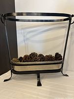 Full view of forged iron frame supporting a long aluminum basket filled with pinecones, showing structure and scale.