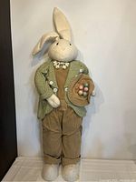 Front view of large stuffed bunny wearing green jacket and corduroy pants holding Easter egg basket.