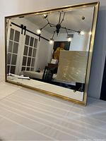 Front view of large beveled mirror with gold metal frame leaning against a wall, showing reflective surface and part of room reflected.