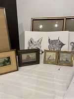 Three outdoor Canada animal prints (fox, lynx, sheep) and vintage sailboat prints, plus four small framed original sailboat paintings and three empty frames, all shown on white fabric surface.