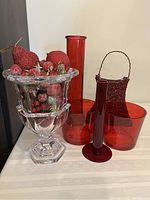 Six-piece grouping of red and clear glass vases, artificial Christmas fruit in clear glass vase, and red sparkling basket with metal handles displayed on white surface.