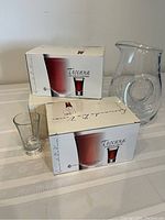 Two boxes of Toscana shot glasses with one shot glass displayed in front, and a clear glass water pitcher with a circular handle hole.