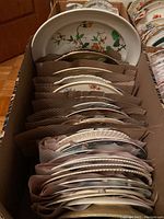 Box containing over 30 various china plates arranged in rows separated by brown paper.