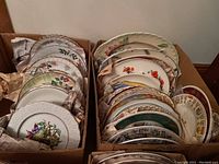 Two cardboard boxes filled with over 30 assorted china plates and serving tray pieces with various floral designs.