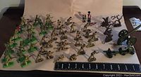 Top view showing the green and beige army action figures in several poses including prone, standing, and running. Also visible is the large artillery cannon with wheels, and brown tree-like props on the right side of the photo.