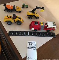 Five metal and plastic construction vehicles with measuring tape for scale showing about 8-9 inches in length of largest vehicles. Items include fire truck, concrete mixer truck, earth movers, and green tractor.