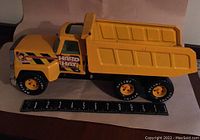 Side view of large yellow and black metal dump truck with 'Hard Hat' decal, six wheels, and detailed features, with ruler for size reference