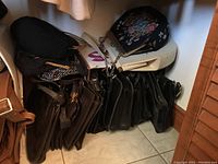 Stack of more than 10 dark colored purses varying from black to brown tones, some leather-like and others fabric, piled together on the floor