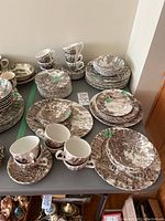 Photo showing multiple stacks of brown transferware plates, bowls, cups, and saucers from the three English patterns arranged on gray table.
